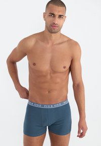 Blaue Baumwollboxer mit einem weichen Bund, der das Logo "PIER ONE" trägt, auf einem Modell vor neutralem Hintergrund präsentiert.