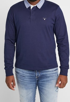 Mann trägt ein marineblaues Langarm-Poloshirt mit hellblauem Kragen und ausgewaschene blaue Jeans, steht vor einem schlichten Hintergrund.