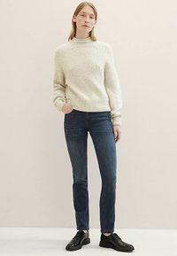 Krém színű kötött pulóver magas nyakrésszel, sötétkék, testhezálló farmerrel párosítva. A modell fekete, fűzős cipőt visel. Egyszínű háttér.