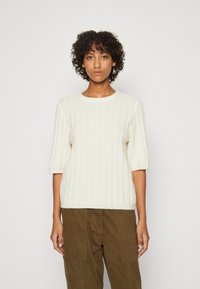 Cremefarbener gerippter Pullover mit kurzen Ärmeln und rundem Ausschnitt, aus weichem Material gefertigt, kombiniert mit olivgrünen Hosen.