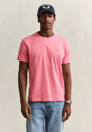 Mann trägt ein pinkes T-Shirt, blaue Jeans, eine marineblaue Kappe mit weißem G und eine Armbanduhr und steht vor einem schlichten hellen Hintergrund.