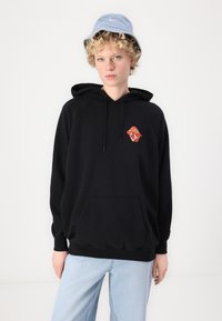 Czarna bluza z kapturem z logo Rolling Stones w kolorze czerwonym i pomarańczowym, z przednią kieszenią i sznurkiem, w połączeniu z jasnoniebieskimi dżinsami i jasnoniebieską czapką typu bucket.