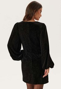 Svart sammet mini-klänning med långa, puffärm och åtsittande midja. Subtil textur med blänkande detaljer. Bakifrån syns en slät silhuett.