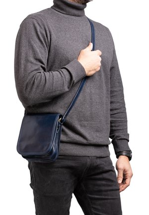 Man draagt een grijze coltrui en zwarte broek, houdt de band van een marineblauwe leren crossbodytas vast.