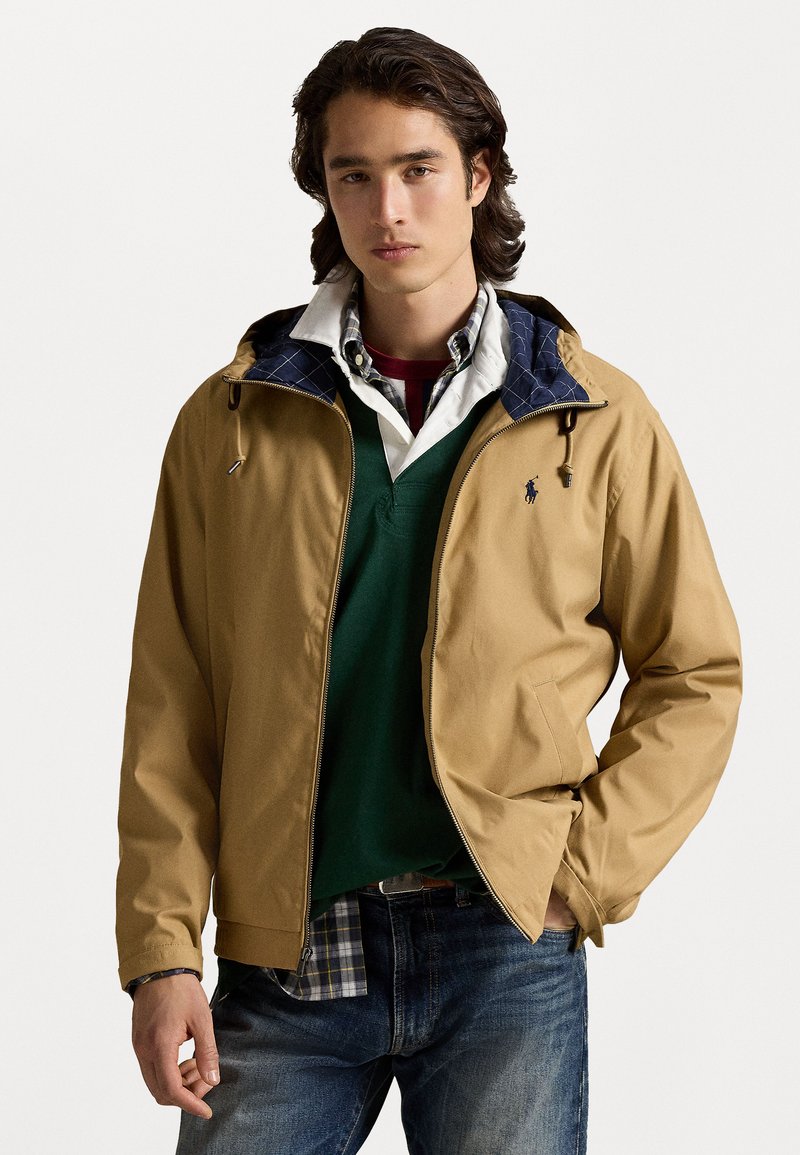 Tan zip-up jacket with a navy quilted lining, layered over a green sweater and a checkered shirt, paired with blue jeans.