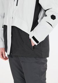 Herrenjacke in Schwarz und Weiß, mit modernem Design, wasserdichtem Material, gewinkeltem Schnitt und hexagonalem Akzent am Ärmel.