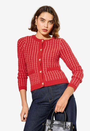 Red knitted cardigan with textured vertical stripes, round neckline, and gold buttons. Paired with dark denim and a black handbag.