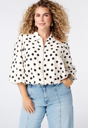 Vrouw met krullend haar draagt een wit zip-up blouse met zwarte stippen en lichtblauwe high-waisted jeans met hand in de zak.