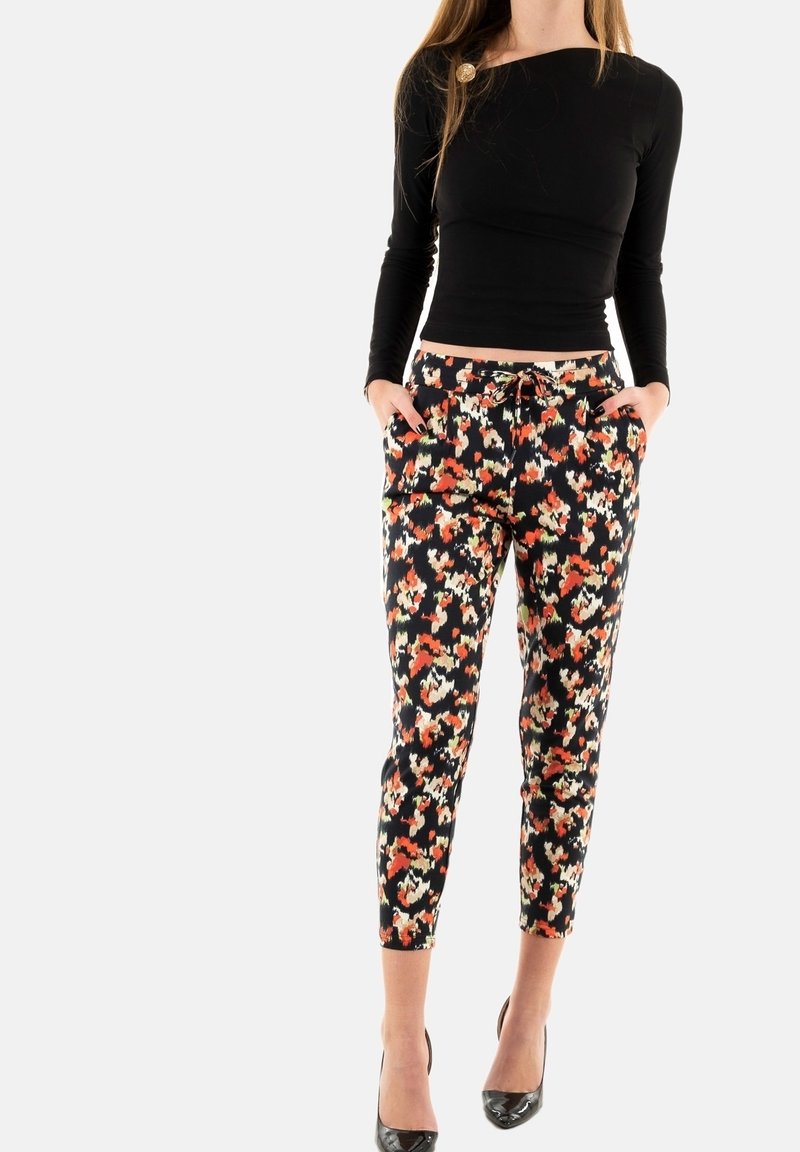 Femme portant un haut noir à manches longues et un pantalon court noir avec un imprimé floral orange, blanc et rouge, debout les mains dans les poches.