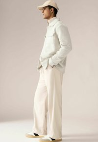 Cămașă lungă cu mânecă, de culoare deschisă și texturată, cu guler, asortată cu pantaloni largi de crem și adidași multicolori. Include o șapcă bej.