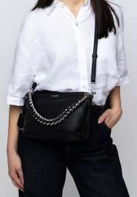 Borsa a tracolla in pelle nera con tracolla in catena argentata, forma strutturata e chiusura con zip. Il modello indossa una camicia bianca con bottoni e jeans scuri.