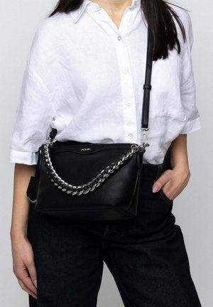 Schwarze Leder-Crossbody-Tasche mit einem silbernen Kettenriemen, strukturierter Form und Reißverschluss-Oben. Das Modell trägt ein weißes Hemd mit Knöpfen und dunkle Jeans.