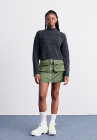 Pull en maille gris, jupe zippée verte avec poches à soufflet, baskets blanches épaisses, chaussettes blanches mi-mollet ; le modèle se tient contre un fond uni.