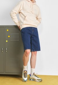 Ljusrosa hoodie med ficka framtill, blå denimshorts och flerfärgade sneakers. Står bredvid ett grått locker med runda gula magneter.