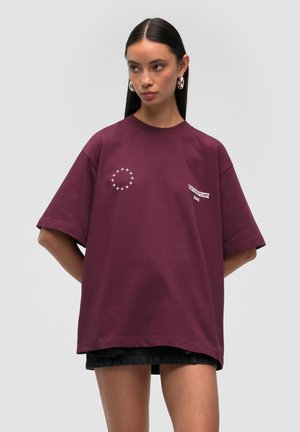 T-shirt oversize bordeaux a maniche corte. Presenta una grafica a forma di stella circolare sul lato sinistro e testo bianco. Materiale in cotone morbido.