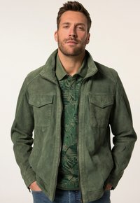 Grüne Wildlederjacke mit Stehkragen, Reißverschluss und zwei Brusttaschen, getragen über einem grün gemusterten Poloshirt. Texturierte Stoffdetails sichtbar.