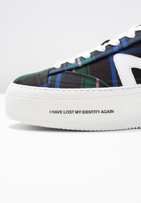 Černá teniska s vícebarevným károvaným vzorem, bílými akcenty a výrazným nápisem na podrážce: "ZNOVU JSEM ZTRATIL SVOU IDENTITU."