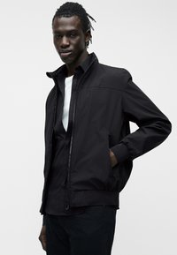 Νεαρός με σκουρόχρωμο δέρμα και dreadlocks, φορώντας μαύρο μπουφάν τύπου bomber πάνω από μαύρο πουκάμισο και λευκό ανδρικό μπλουζάκι, ποζάρει με το χέρι στην τσέπη του μπουφάν.