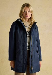 Námornícky modrý parka kabát s kapucňou, predným zipsom, dvoma bočnými vreckami a sťahovacou šnúrkou. Nosený cez vzorovaný sveter. Pohodlný strih.