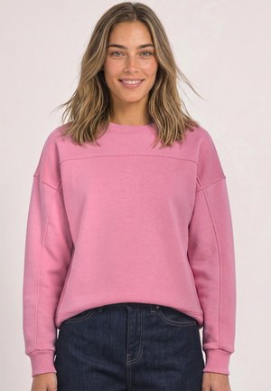 Femme aux cheveux blonds mi-longs souriante, portant un sweat-shirt rose à manches longues et un jean bleu foncé, sur un fond clair uni.