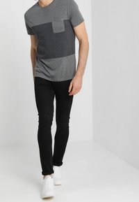 Camiseta gris de manga corta con una franja horizontal gris oscuro y un bolsillo, combinada con pantalones negros ajustados y zapatillas blancas.