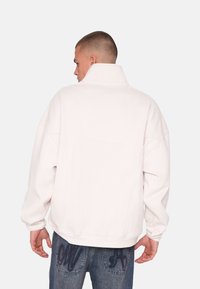 Hvid oversized fleecejakke med høj krave og synlige sømdetaljer, parret med mørke jeans og detaljeret syning på baglommerne.