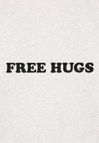 T-shirt gris avec un texte brodé noir en gras qui indique "FREE HUGS" dans une police épaisse et arrondie. Tissu doux et texturé.