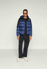 Moteris, dėvinti mėlyną ir juodą siuvinėtą striukę, juodas krovinių kelnes, béžinius sportinius batelius ir baltus saulės akinius, stovi prieš paprastą šviesų foną.