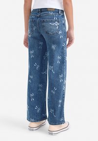 Blauwe denim jeans met wijde pijpen, voorzien van witte strikpatronen, een vijfzakkenontwerp en rafelige zoom, gecombineerd met lichtgekleurde sneakers.