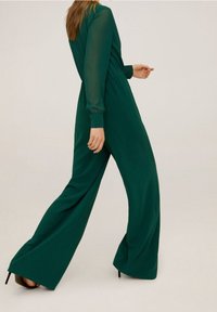 Jumpsuit vert avec une texture lisse, des manches longues et une taille cintrée. Design à jambes larges et légère brillance, associé à des talons hauts.