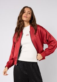 Czerwona satynowa kurtka bomberka z zamkiem z przodu i ściągaczami przy mankietach, noszona na dopasowanym białym topie z czerwonym haftowanym napisem. Czarne spodnie w wzory.