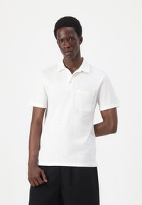 Weiße kurzärmelige Poloshirt mit strukturierter Oberfläche, einem spitzen Kragen und einer Brusttasche, kombiniert mit schwarzen Hosen.