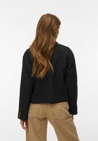 Femme aux longs cheveux bruns ondulés portant une veste gris foncé et un pantalon beige, debout de dos sur un fond uni.