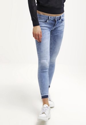 Osoba ubrana w jasnoniebieskie dopasowane dżinsy i białe trampki, stojąca z jedną nogą lekko do przodu na jednolitym białym tle.