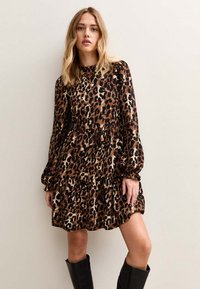 Leopardmönstrad klänning med långa ärmar, markerad midja och lager-på-lager-kjol. Tillverkad i mjukt tyg, med svart- och bruna toner. Svarta knähöga stövlar.