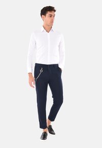 Camicia bianca a maniche lunghe con bottoni, pantaloni blu scuro con un accento in catena e mocassini neri. I pantaloni sono affusolati e hanno una texture liscia.