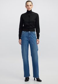 Schwarzes, langärmliges Oberteil mit Knöpfen und leichtem Schimmer, kombiniert mit hellblauen, weiten Jeans. Modell trägt schwarze, hohe Stiefel.
