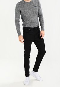 Musta slim-fit farkut, joissa on sileä pinta, yhdistettynä harmaanhehkuiseen pitkähiharaiseen paitaan. Valkoiset lenkkarit täydentävät asukokonaisuuden.