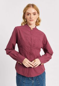 Donna che indossa una blusa prugna aderente con bottoni dorati, abbinata a una gonna di jeans blu, in piedi davanti a uno sfondo bianco.