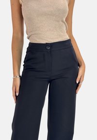 Zwarte wijde broek met een textuurafwerking, voorzien van een sluiting met een knoop aan de voorkant en zijzakken, gecombineerd met een beige geribbelde tanktop.