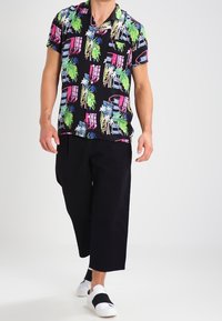 Camiseta negra de manga corta con patrones coloridos de palmeras y ventanas, combinada con pantalones negros holgados y zapatos blancos sin cordones.