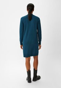Abito maglione verde acqua con maniche lunghe, polsini e orlo a coste, realizzato in un tessuto morbido. Indossato con stivali alla caviglia neri, visto da dietro.