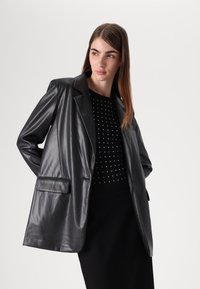 Blazer in finta pelle nera con una tasca sul petto, taglio rilassato e rever affilati, indossato sopra un top e una gonna nera con borchie.