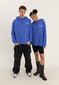 Mladý muž a žena stojící vedle sebe, oba v odpovídajících modrých mikinách s kapucí, černých kalhotách a obuvi na neutrálním pozadí.