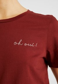 Burgunderfarvet bomulds T-shirt med korte ærmer og rund hals. Har hvid broderet tekst "oh oui!" på venstre brystområde.