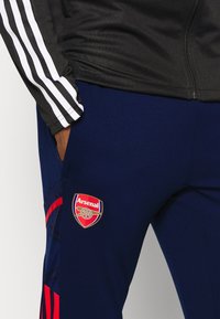 Černá sportovní bunda s bílými pruhy, spárovaná s námořnickými tepláky s logem Arsenalu a červenými akcenty na boku. Hladká textura.