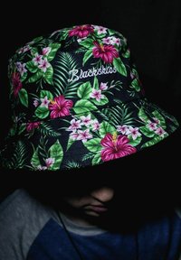 Cappello a secchiello nero con un vivace motivo floreale che presenta hibiscus rosa e foglie verdi, con il marchio "Blackskis" ricamato in bianco.