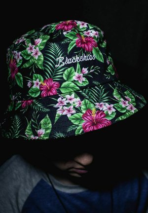 Melna burbuļvāciņš ar dzīvīgu ziedu rakstu, kas sastāv no rozā hibiskiem un zaļām lapām, ar baltu izšūtu zīmolu "Blackskis".