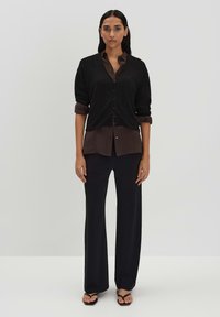 Schwarzes geripptes Hemd mit Knöpfen, über einer dunkelbraunen Bluse getragen, kombiniert mit lockeren schwarzen Hosen und schwarzen Sandalen.