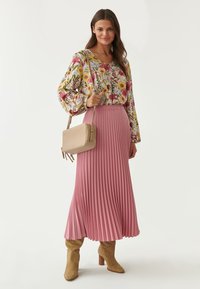 Blomstret bluse med ballonærmer, parret med en plisseret lyserød nederdel. Tilbehør inkluderer en beige crossbody taske og brune knæhøje støvler.